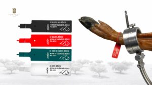 Los cuatro precintos oficiales del jamón ibérico —negro, rojo, verde y blanco— junto a una pieza en malla negra RH Gourmet sobre mármol