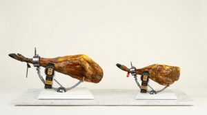 Jamón ibérico y paleta ibérica de RH Gourmet en jamonero, lado a lado, mostrando la diferencia de tamaño y forma