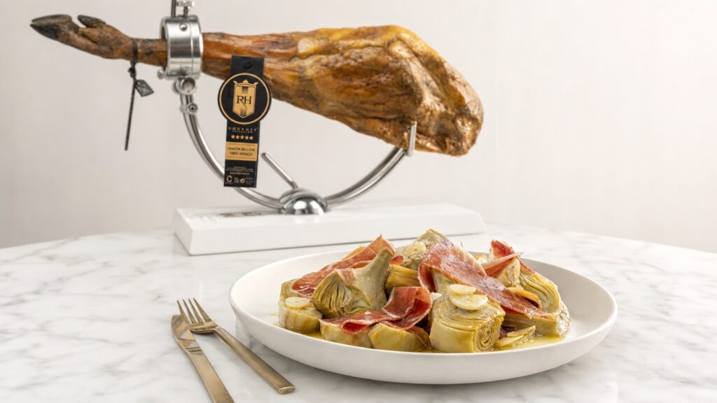 Plato de alcachofas con lonchas finas de jamón ibérico y una pieza de jamón RH Gourmet al fondo en jamonero blanco.