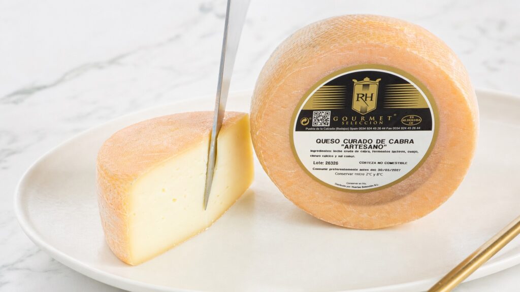 Cuña de queso curado de cabra RH cortándose junto a la rueda entera sobre mármol blanco
